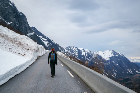 Young woman hiker with backpack walking up on a mountain road with large snowdrifts, springtime, back view.の写真素材