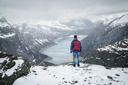 Man traveller with backpack standing back on edge and looking at scenery mountain landscape under snow, Trolltunga, Norway in may.の写真素材