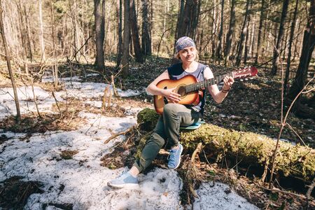 Young woman playing guitar sitting on old tree covered with moss at sunny spring forestの写真素材