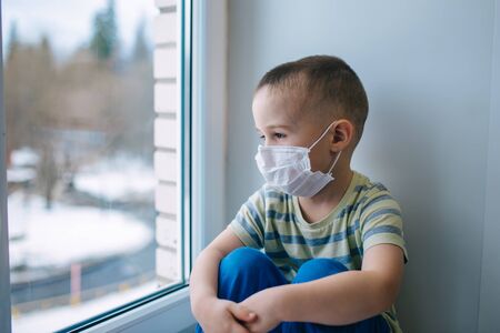 Sad little boy in medicine mask sitting near window and looking outside, quarantine at home, virus protectionの写真素材