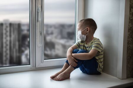 Sad little boy in medicine mask sitting near window and looking outside, quarantine at home, virus protectionの写真素材
