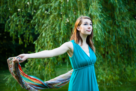 Beautiful girl is slowly spinning around in front of a willow tree. Carefree, happy and joyful in a park during summer.の写真素材