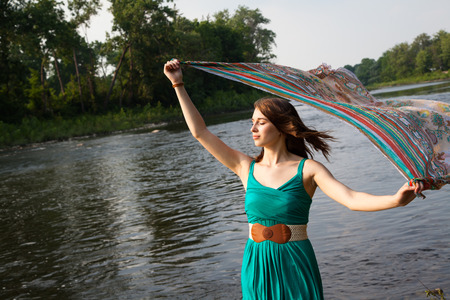 Young woman with a teal green dress stands near a river and lake holding teal colorful scarf in the wind.   She is relaxed and serene while feeling the summer breeze.の写真素材