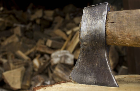 Closeup of an axe sticking in a chunk of firewood in front of a staple of firewood.