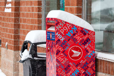 A Canada Post street letter box stands outside shops in a small outdoor shopping plaza during a winter snowfall, with a layer of snow on top of the mailbox.のeditorial素材