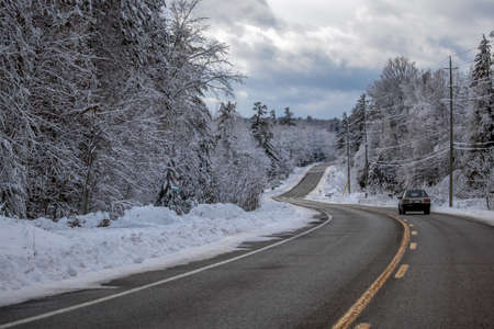 An older car is driving down a winding country road in the winter, with forest trees on either side...