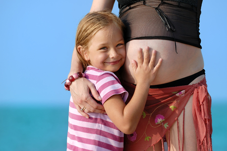 Little girl hugging her pregnant mom on the beach, sea in backgroundの写真素材