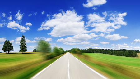 Road among green fields with motion blur effect, blue sky and white clouds in the background