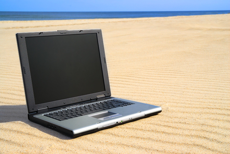 Modern laptop lying on sand. Beach and clean blue sky in background.の写真素材