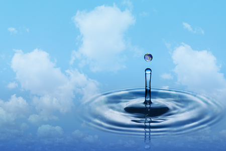 Drop of rain with earth image falling on smooth water surface. Blue sky and white clouds in background.の写真素材