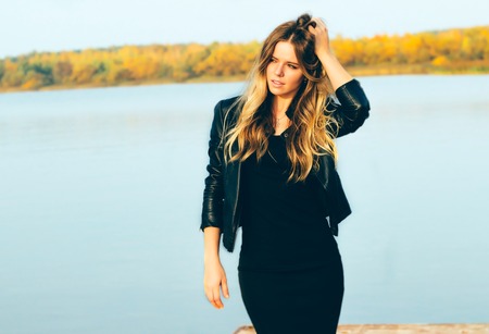 Young beautiful blonde woman in autumn park with lake in dark leather jacket smile with perfect...