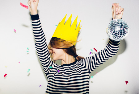 Birthday party, new year carnival. Young smiling woman on white background celebrating brightful event, wears stripped dress and yellow crown. Sparkling confetti, having fun, dancing, laugh, smile.の写真素材