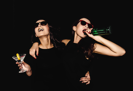 Two beautiful women black night fashion dress posing isolated on a black background. Pretty brunette girl friends twins having fun drinking cocktails. Singing and dancing. Bottle glass of alcohol.の写真素材