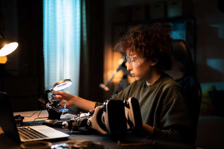 Soldering in the evening. A child is sitting at desk on chair next to lighted lamp, boy is soldering...