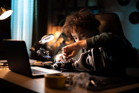 A young DIYer builds a robot. Child sits at desk and connects cables, toy electronics, solders wires, boy is interested in roboticsの写真素材