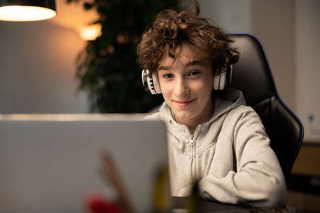 A young boy with curly hair is happy with remote learning. A student sits in front of a computer with wireless headphones listening to a teacher lecture. The lessons take place over the Internet.の写真素材