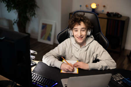 A boy sits at a desk at home in front of a laptop. The child is listening to music on wireless...