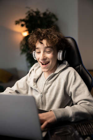 A child uses a laptop at desk. The boy with curly hair is wearing headphones is listening to music, playing computer games, browsing the internet, chatting with friendsの写真素材