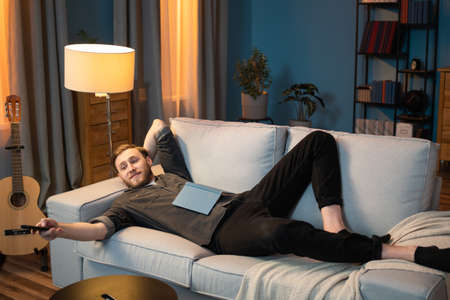 Adult man is resting after work in the evening in living room, lying along the couch, propped up by head, leg bent at the knee, a book lying on stomach, switching channels with the TV remote controlの写真素材