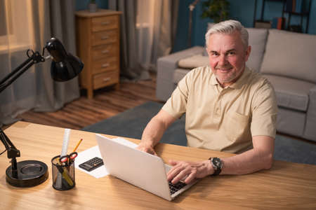 An elderly smiling man works on his laptop in the evening at his desk and lamp. Mature businessman happy to work online. The man in the yellow T-shirt and wristwatch is working remotely.の写真素材