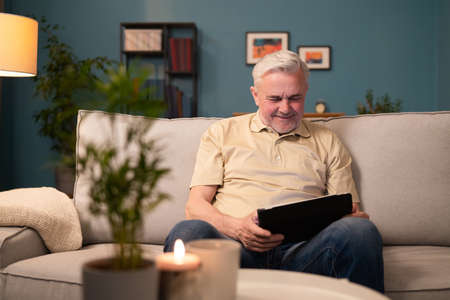 Smiling grey-haired man relaxing in living room with tablet. A pensioner reads the news on the internet. Senior man reads ebooks, online blogs. A senior man learns to use electronic devices.の写真素材