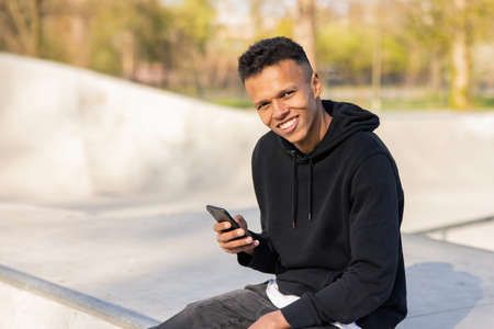 A dark-skinned boy is waiting in the park, holding his phone in his hand to write a message, while...