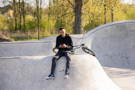 The student spends his free time being active by riding his bike, bmx on ramps in the park. He is sitting on a concrete hill with his feet loosely down and holding his phone with a wide smileの写真素材