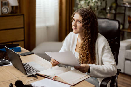 The girl teenager in a white sweater, studies online from home, sits in an office chair, summarizes...