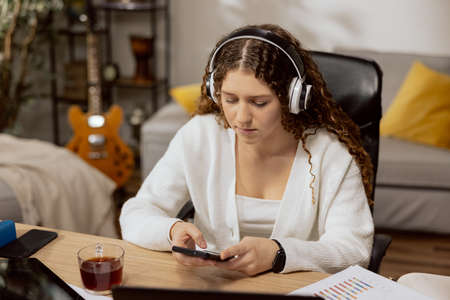 Conceived woman with curly hair, in wireless headphones sitting in the living room, holding a phone, surfing the Internetの写真素材
