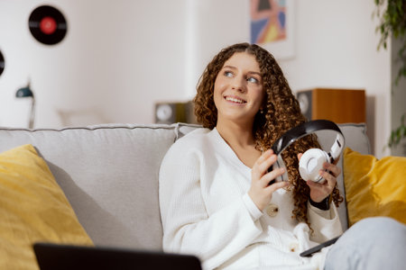 Brunette with curly hair and snow-white smile. The girl is wearing sweater and blue jeans, holding wireless headphones and a phone on knees. Dreamy woman looks up at the mountain, happy.の写真素材