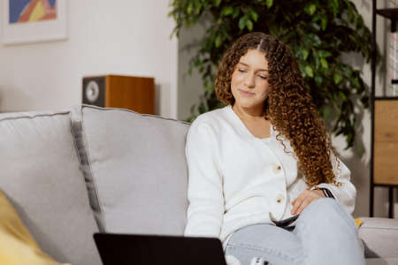 Young, beautiful, smiling girl with long, curly, brown hair, sitting on a gray sofa in the living room. Woman watching favorite TV series through laptop.の写真素材
