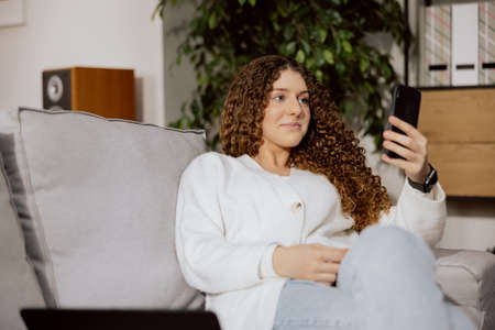 Young, beautiful woman, corporate worker, working remotely from home. The girl sits on a gray, comfortable sofa, holds a phone in his left hand, communicates with colleagues, smilesの写真素材