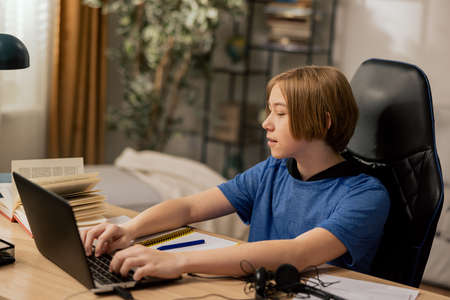 A young boy uses laptop while sitting in room on a chair at desk. The child is doing homework, writing a paper, taking notes on school reading, remote teaching.の写真素材
