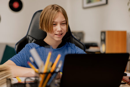A boy with longer hair sits in front of laptop in room doing homework, watching science videos on...