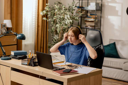 A young boy listens to music from his phone on headphones while studying. Child sits at home office at desk in front of laptop, studying for test, doing homework.の写真素材