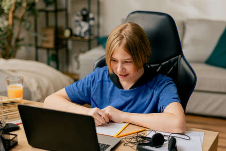 School age boy with long hair spends time in room at desk on chair doing homework in front of computer, remote teaching, e-learning tutoring individual with teacher online.の写真素材