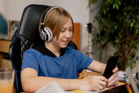Young boy with wireless headphones on ears sits in chair at desk with a phone in hand. The teenager is listening to music, watching videos, sending messages to friends.の写真素材