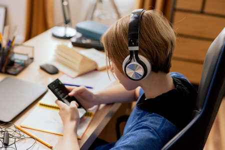 A teenager listens to music on wireless headphones while doing homework. A young boy searches for a study playlist on his phone and uses an app with songs.の写真素材