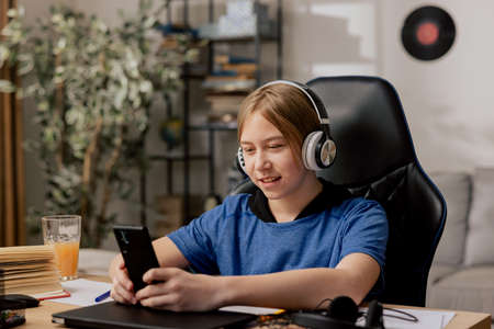 Young boy sits in room in morning drinking orange juice, holding phone in hands, watching videos, listening to music on wireless headphones, entertainment before homework.の写真素材