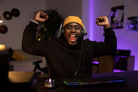 A man wins in a computer game. The boy in the headset raises his hands in the air screaming with the joy of victory. Dark room at night lit by led, neon lights.の写真素材