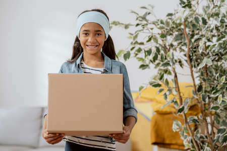 Sweet girl helps her parents during the move, carries things from room packed in cardboard box,...