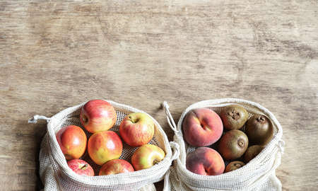 Organic fresh fruits - apples, kiwi, peaches in reusable eco friendly natural bags on the wooden background. Package-free food shopping. Zero waste concept. Reuse, reduce, refuse. Top view, copy spaceの写真素材