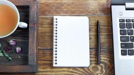 Wooden tray with a cup of herbal tea, a laptop and an opened notebook and a black pen on a dark...