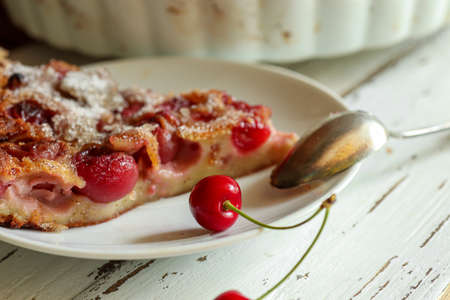 A slice of homemade clafoutis dessert - traditional french cherry pie, fresh cherry berries and a teaspoon on a white plate on a white wooden table, close upの写真素材