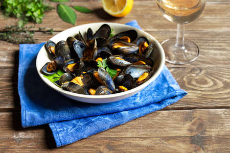 Delicious fresh steamed mussels in white wine sauce in a white plate, a lemon, parsley and a glass of wine on a wooden tableの写真素材