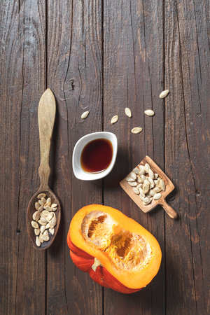 A white bowl with pumpkin seed oil, pumkin seeds in wooden spoons and fresh autumn pumkin on an old dark wooden table, top vertical view with copy spaceの写真素材