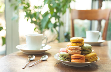 Multicolored macarons or macaroons, traditional French dessert on a plate on a round wooden table with white cups, tasty breakfastの写真素材