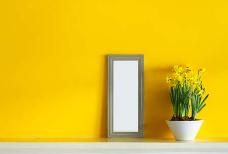 A white ceramic pot with blooming yellow daffodils and a gray photoframe on a yellow background with...