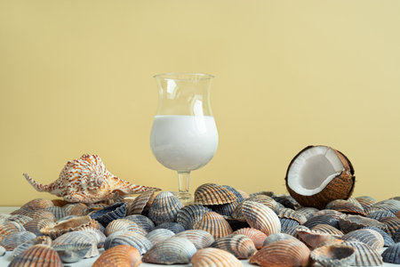 A glass of coconut milk cocktail, a coconut and seashells on a light yellow background, summer organic beverageの写真素材