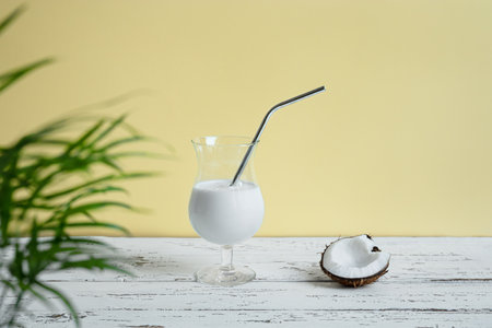 A glass of coconut milkshake with reusable metal cocktail tube and a piece of coconut on a white wooden tableの写真素材